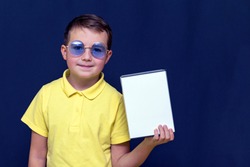 Happy caucasian boy wears yellow t-shirt with blue glasses and holds white box with a layout on a blue background with copy space.