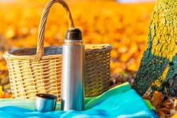 A wicker basket metal steel thermos and a cup with a hot drink, tea, coffee stands on a autumn leaves background in nature picnic autumn forest.Selective focus,blured background.