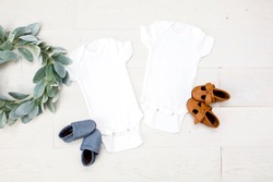 Two blank white baby bodysuits on white wood background with baby shoes and green wreath, newborn twin bodysuit mockup