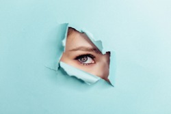 Eye of the beautiful woman looking through hole in blue paper. Spy eye watching through hole. Copy space.