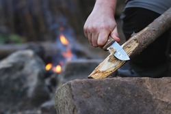Close up of the hands of a man carving off timber to lit a fire, camp fire on the background. Hand holding knife cutting a wooden stick. Bushcraft and outdoors survival activities concept. 