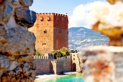 Beautiful view of ancient Red Tower in Alanya, Turkey, between two merlons of old stone wall. Sandy beach and turquoise sea under old historic walls on sunny day. Blurred foreground. Selective focus.