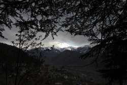 Evening silhouettes of pine trees against the background of an Indian mountain village at the foot of mountain peaks.
