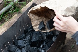 Black charcoal as natural fuel for warming and cooking. Hand holds paper bag with coal to adding it into brazier.