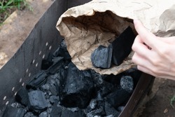 Black coal as natural fuel for cooking barbeque in brazier. Hand holds paper bag with charcoal 
