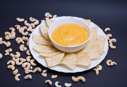 Vegan cheddar cheese in a bowl and nachos to dip in a plate on a black background with cashew nuts.