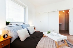 Simple bedroom in apartment with walk-in closet and small bathroom hidden behind sliding door. In addition to bed, there are bedside tables, lamps, two windows and a tray with cups on the bed.