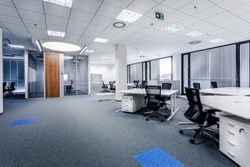 Part of ordinary office room decorated in modern style with meeting room,large windows with blinds,carpeting,ventilation,escape signs,white furniture (tables, shelves, drawers) and dark office chairs.
