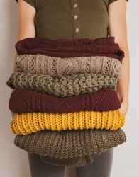 A young woman holds a stack of warm clothes in autumn colors. Sweaters in yellow, dark green, brown