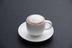 White cup of cappuccino on a black textured stone slab, side view. White foam.