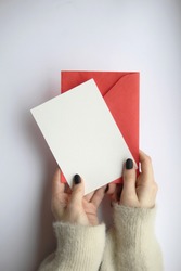 female hands in a sweater with manicure, holding a white sheet and a red envelope on a white background. postcard layout