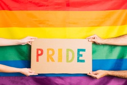 hands of man and woman holding a cardboard with the word pride written in colors on the background gay pride flag