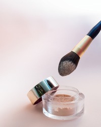 A golden makeup brush is dipped in a glass jar with powder on a pink background