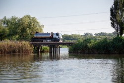 Silver stainless steel tanker truck on concrete bridge over a small river with green reeds. Road bridge with concrete columns.
