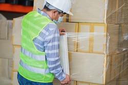 close up view of man worker wearing protective face mask and safety suite wrapping stretch film parcel on pallet in factory warehouse, logistic industry concept. 