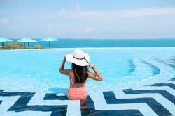 Carefree Woman in bikini and a straw hat relaxing in infinity swimming pool looking at  seaview. Luxury resort. Beautiful destination summer vacations. Back view