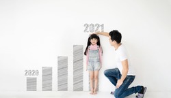 Dad measures height increase of her child daughter at white brick wall with graph growing growth year 2020 to 2021