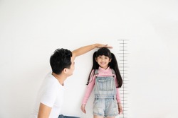 Dad measures height increase of her child daughter at white brick wall with scale