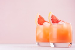 Cold grapefruit cocktail in two glasses with ice and pieces grapefruit on pastel pink background. Fresh summer healthy diet beverage.