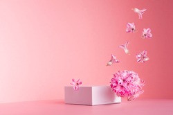 White square podium for display cosmetic and goods with hovering flow of fresh spring flowers  on gentle pastel pink background, copy space, modern fashion style.