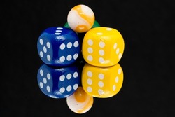 A macro closeup image of a blue and a yellow dice next to each other with an orange and white marble balance on top.  All against a black background reflected in a mirror.
