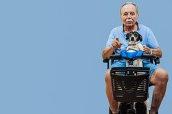 Happy elderly disabled person or senior with a dog on electric wheelchair or a mobility scooter. Modern technology for people disabilities. Pale blue background. Copyspace.