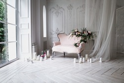 vintage sofa of soft pink color, decorated with flowers and greens, stands in a classic room on a white wooden floor surrounded by lighted candles in glass candlesticks near  large window and curtains