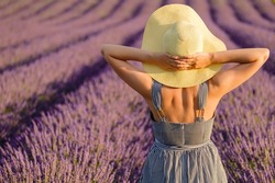Woman in sundress puts hands on straw hat standing on lavender field. Young lady enjoys beautiful nature on sunny summer day backside view