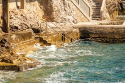 Metal ladder for swimmers descending into deep water on narrow rocky beach. Blue sea waves wash coastline with old stone steps leading to resort