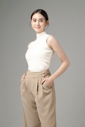Confident Asian woman puts her hands in pocket and looks at the camera isolated over grey background. Portrait of pretty girl in studio. Perfect body.