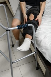 Woman wearing adjustable knee brace after leg fracture sitting on bed trying to stand up with walker. Recovery and rehabilitation after surgery and installation of plates. Selective focus