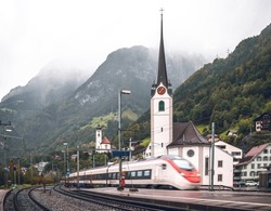 High-speed Swiss railways train arriving at Flüelen station, passing church and rainy Swiss Alps. Mountainous landscape of Switzerland. 
