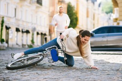 Fall with bike. Young unhappy woman on a city street falling with a bicycle on the road and a man running hurrying behind