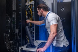 Server checking. Young bearded man in tshirt and jeans with badge in server room near open cabinet checking