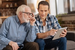 Look here. Pleasant young man pointing at the tablet, showing an interesting article to his elderly father while the man paying attention to him