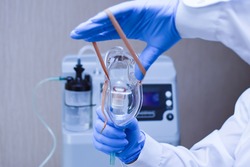 Hands in medical gloves hold out an oxygen mask against the background of the oxygen generator concentrator  to deliver at low lung oxygen saturation for Covid-19 pneumonia. Focus on oxygen mask
