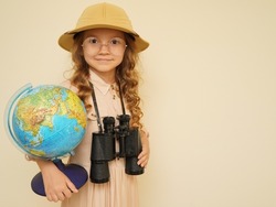 Happy child plays the discoverer. Unique child. Travel, adventure, vacation and discovery concept. A kid in a traveler's hat with binoculars and a globe in his hands on the beige background. 