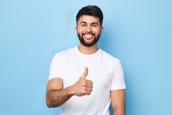 Happy bearded man in stylish white T-shirt with beaming smile showing thumb up, isolated over blue background, studio shot. good job, well done. agreement concept