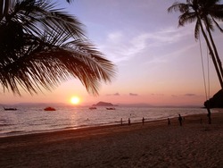 Sunset on the beach with palm tree leaf, palm trees and swing silhouette with clouds in the sky, people on the beach and boats on the horizon. Sunset scenery on tropical island in Thailand.