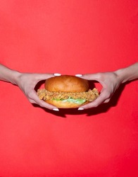 A woman wearing white nail polishe is holding a crunchy chicken burger in her hands in the middle of a red background.