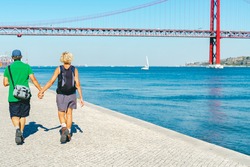 happy retired couple of woman and man with bag pack walking exploring Lisbon, enjoying view on Red bridge Ponte 25 de Abril. Suspension Bridge over the Tagus river. elderly pensioner family traveling