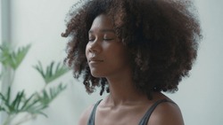 African young adult female engages in a deep meditation or yoga practice at home. With her eyes gently closed, she finds inner peace and calm amidst the demands of daily life.