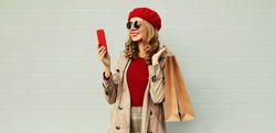 Autumn portrait of smiling woman with smartphone and shopping bag wearing a coat, red french beret over gray background