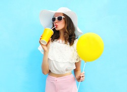 Fashion pretty young woman wearing a straw hat, sunglasses with air balloon drinks fruit juice from cup over colorful blue background