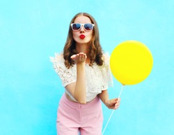 Pretty young woman in sunglasses with air balloon sends an air kiss over colorful blue background