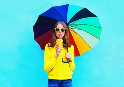 Fashion pretty woman with fresh fruit juice cup and colorful umbrella in autumn day over blue background wearing a yellow knitted sweater