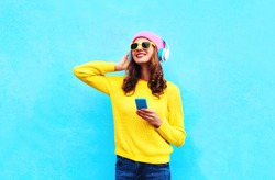 Fashion pretty sweet carefree woman listening to music in headphones with smartphone wearing a colorful pink hat yellow sunglasses sweater over blue background