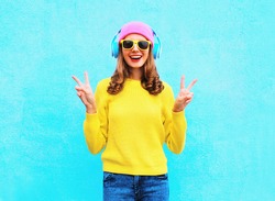 Fashion pretty cool smiling girl in headphones listening to music wearing a colorful pink hat yellow sunglasses and sweater over blue background
