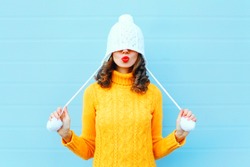 Happy cool girl blowing red lips makes air kiss wearing a knitted hat, yellow sweater over blue background
