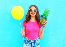 Fashion pretty smiling woman with yellow air balloon and pineapple wearing a pink t-shirt over colorful blue background